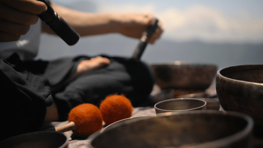 Person in nature playing a Tibetan singing bowl surrounded by multiple bowls, deepening meditation through sound and resonance