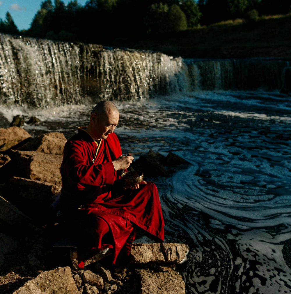 Monk playing a Tibetan singing bowl beside flowing waters in nature, embodying the Himalayan craft tradition behind Serene Streams